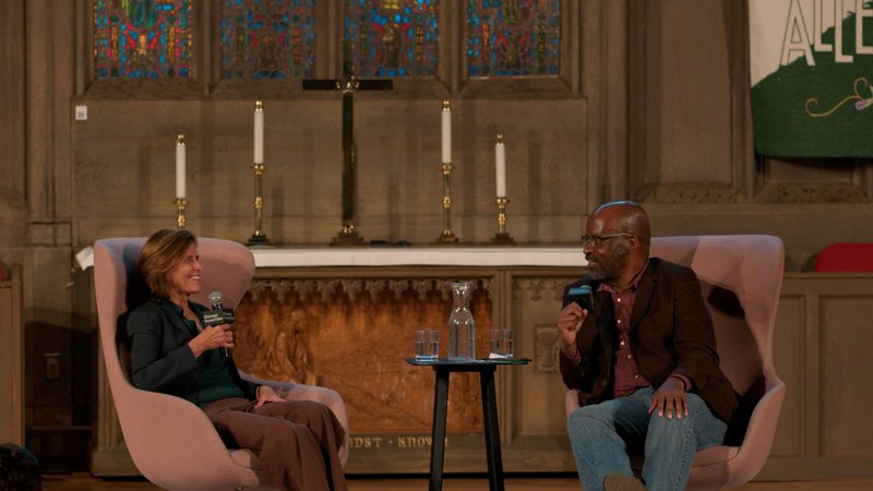 A close up of two people on stage at the front of a cathedral, sitting in large pink chairs. To the left, a white woman with chic chin-length hair smiles. To the right, a Black man leans forward and talks into a handheld mic, wearing a brown suit coat and pants.