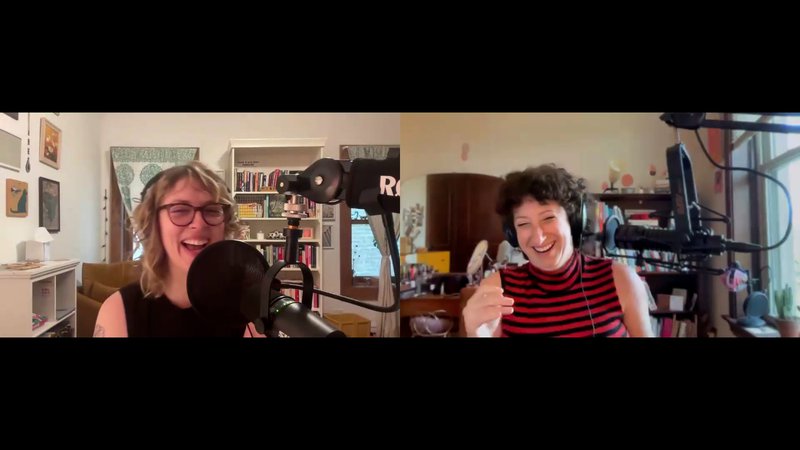 A screengrab of two white women laughing on a Zoom call. In different rooms, they’re both in front of bookcases and speak into professional microphones. The woman on the left has blond hair and glasses, the woman on the right has brown curly hair and wears a black and red striped turtleneck tank.