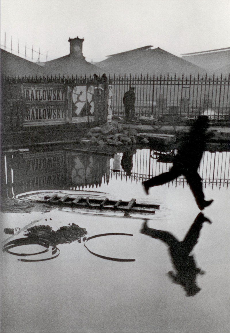 alt text: A dreamy greyscale photo of a man jumping over a puddle. His reflection is perfectly captured so he appears to be levitating.