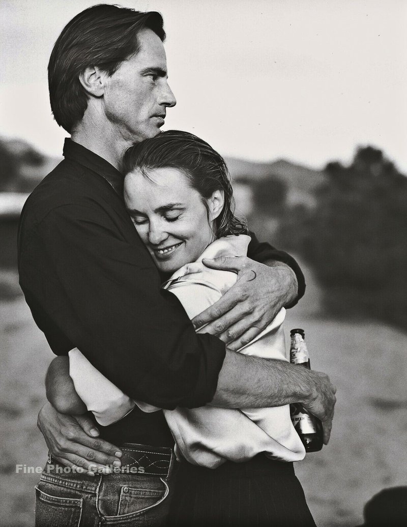 alt text: A black and white photo of a serious man lovingly embracing a woman in a white shirt. She smiles into his chest while he holds a beer.