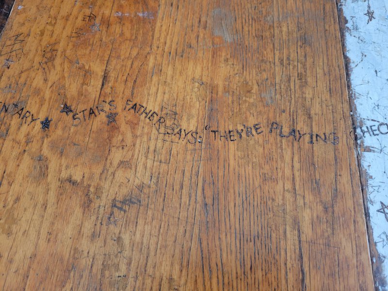 A photo of a closeup of the top of a wooden table, with stars and words etched into it in a squiggly line reading “...STARS FATHER SAYS “THEY’RE PLAYING CHECK…” / Una foto de un primer plano de la parte superior de una mesa de madera, con estrellas y palabras grabadas en una línea ondulada que dice "... EL PADRE DE LAS ESTRELLAS DICE "ESTÁN JUGANDO JAQUE..."