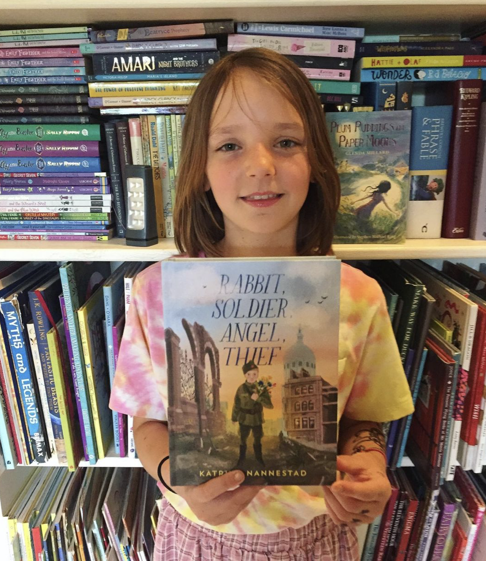 A ten year old girl stands in front of a book shelf overflowing with books and is holding a copy of "Rabbit, Solider, Angel, Thief"