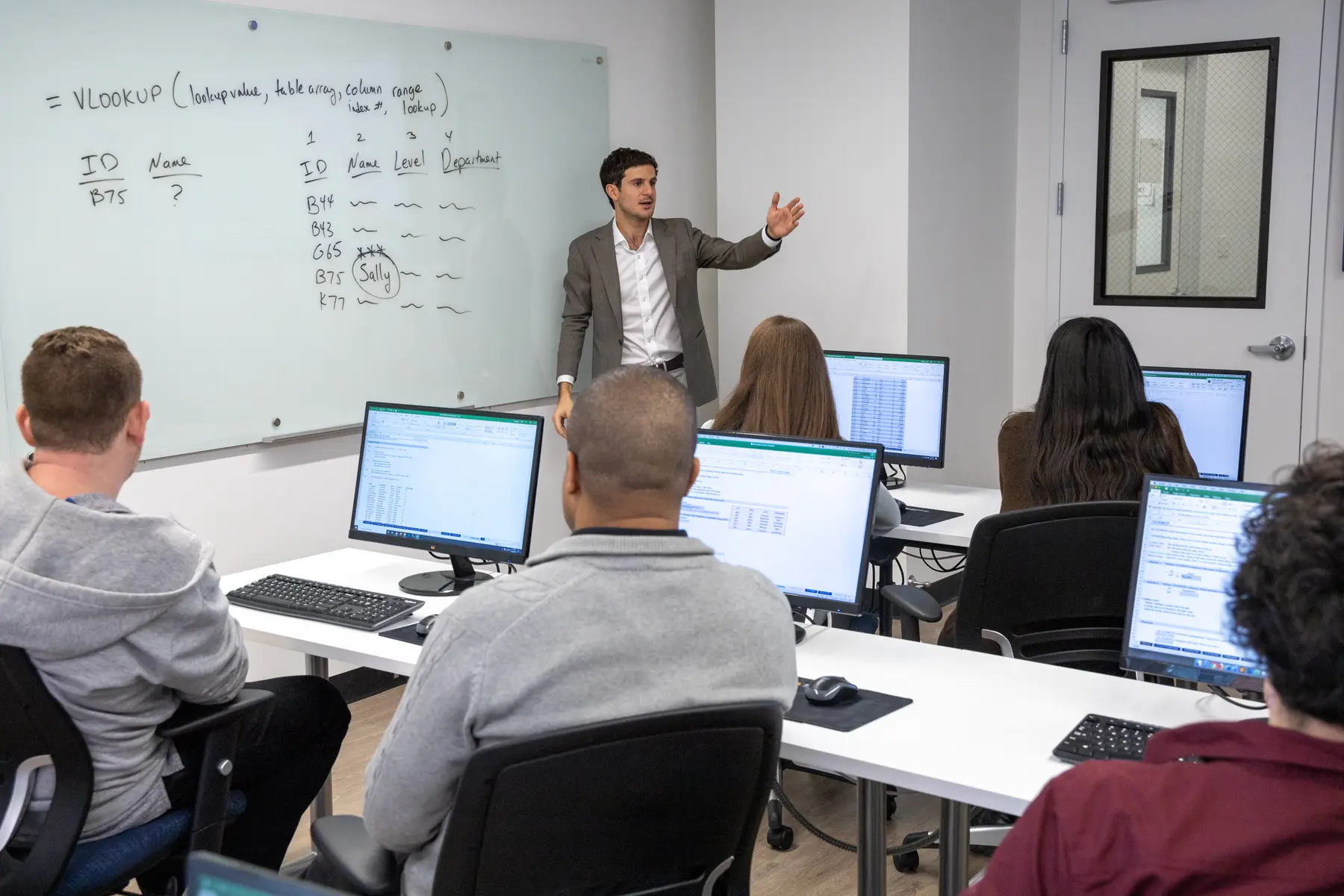 A classroom at Noble Desktop in New York City, where an instructor is teaching Data Analytics Technologies. The students, seated at computers, are engaged in a live or live online session while the instructor stands at the front of the room.