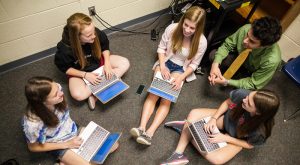 Students with their teacher seated on the floor with their laptop