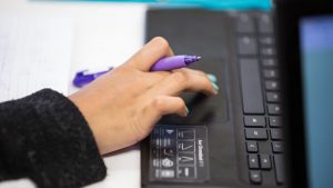 Student's hand holding a pen while typing on the keyboard of a laptop