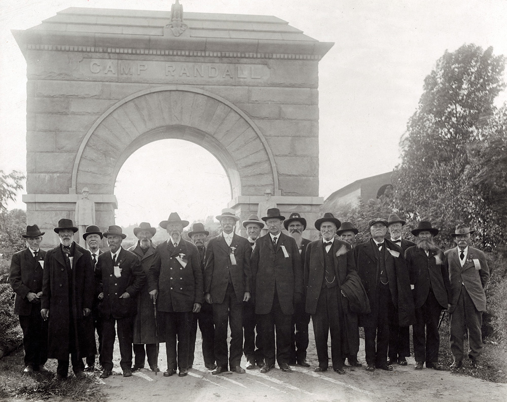 Group portrait photograph of Civil War Veterans standing in front of the Camp Randall Memorial Arch, located in Madison, Wisconsin The veterans are dressed in civilian suits, wearing ribbons and badges, likely from the Grand Army of the Republic.