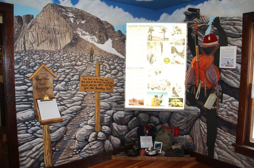 Long's Peak and a hiker are painted on the wall at the left of the image in a museum exhibition on hiking and climbing. There is a trail register and sign in front of the peak. On the right side next to the informational display, there is a climber with wearing a red hat carrying a red backpack with ropes, water bottle, and other climbing gear. 