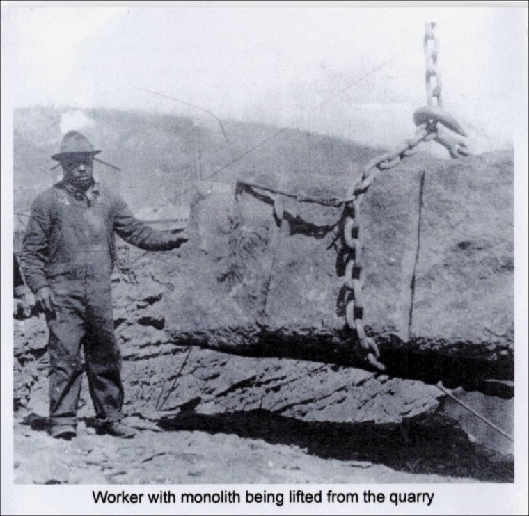 An early 20th century worker stands beside a massive brownstone obelisk.