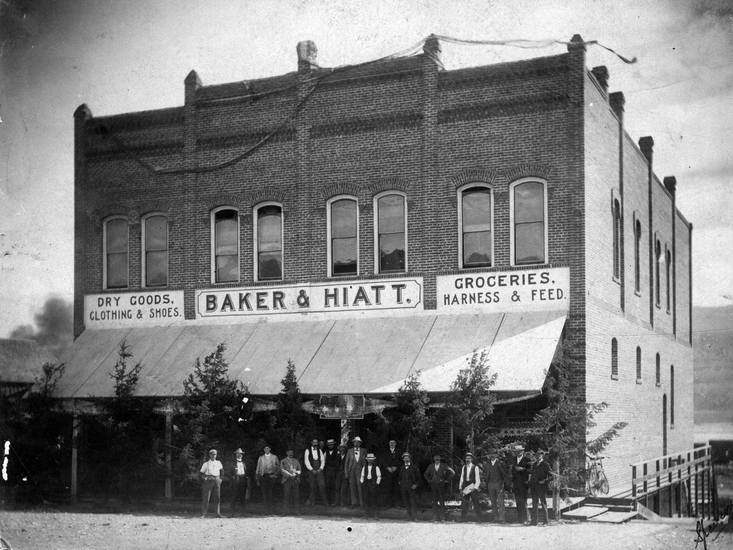 Black and White Photo of Baker and Hiatt building, 23 South Wenatchee Avenue, fourth brick building constructed at in the relocated town site of Wenatchee.  Opened for business in 1897 with W. T. Rarey as manager. James Edward Ferguson was his one and only clerk. Others in the picture are Robert A. Farmer, head of the United States Geological Survey, and his crew. They mapped this section, establishing the  elevations in the Lake Chelan and Methow Valleys, Waterville, Wenatchee and Lake Wenatchee sections. Ed Ferguson, third from left, was a member of the crew. It is noted that the evergreen trees may have been brought in as Christmas decorations.