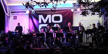 A group of musicians play a variety of instruments on a stage in front of a MoCA banner.
