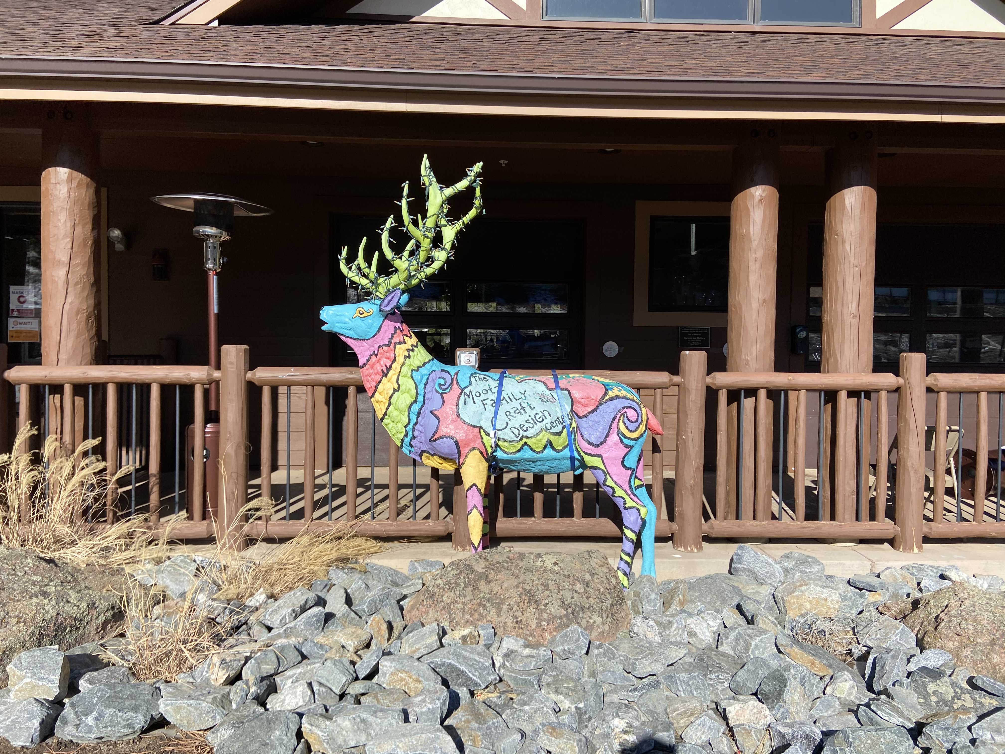 This sculpture of a male elk is painted in various stripes and swirls of bright blue, pink, red, yellow, lime green, and purple. His antlers are lime green and wrapped with lights.