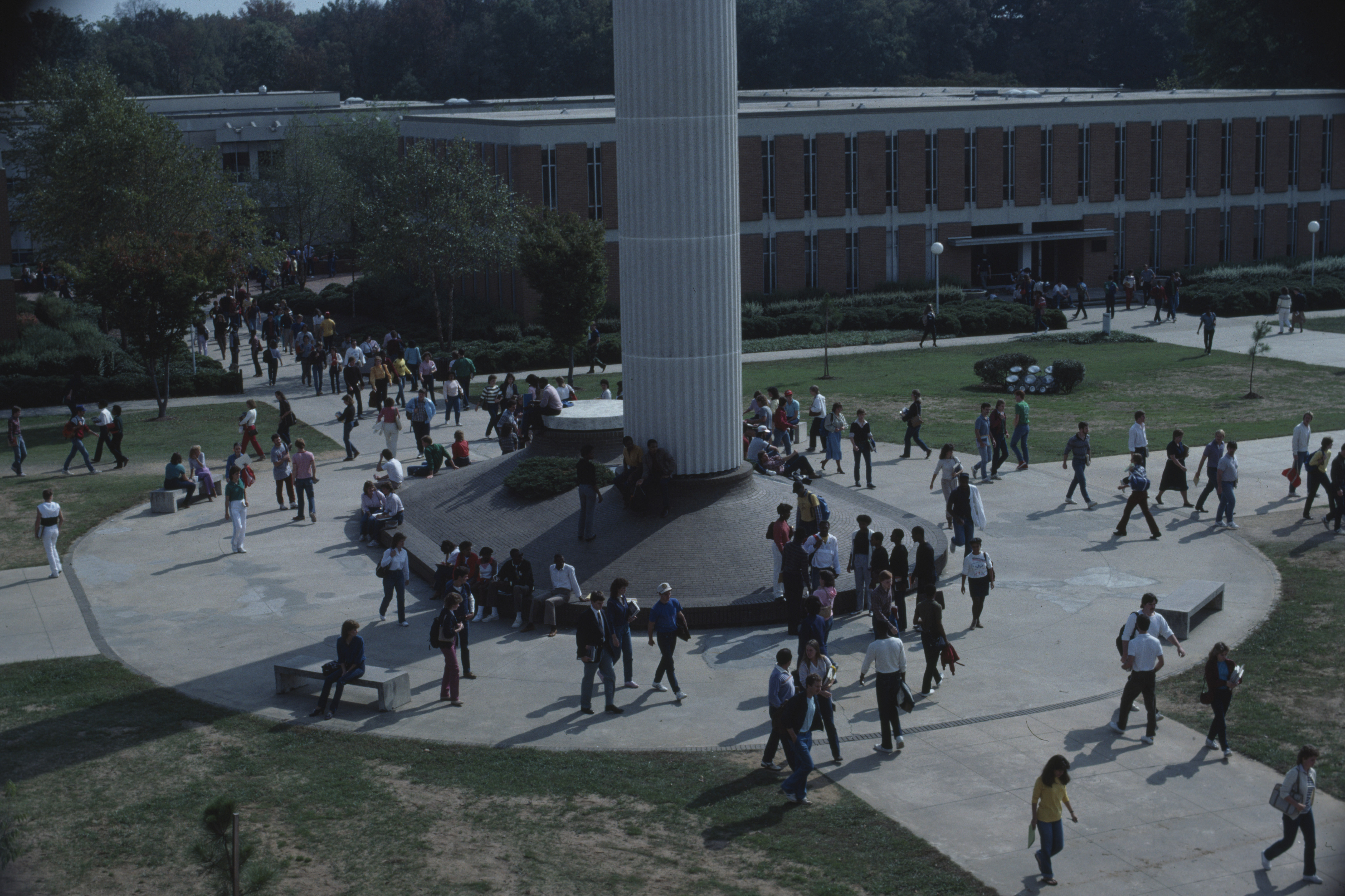 Elevated view of the base of the Belk Bell Tower from around the 1970s-1980s. Many students walk on the path around the base, and a building can be seen in the background.