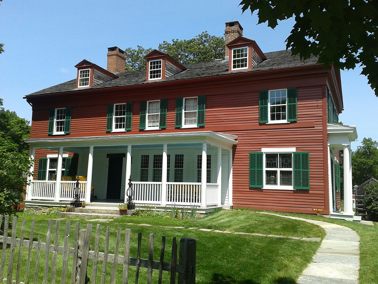 The front of the Weir House. The two-story home features reddish brown siding and green shutters. There is a white covered porch along about one third of the house. Small chimneys and dormers are featured on the sloped, black roof. A flat front yard is enclosed with a wooden fence.