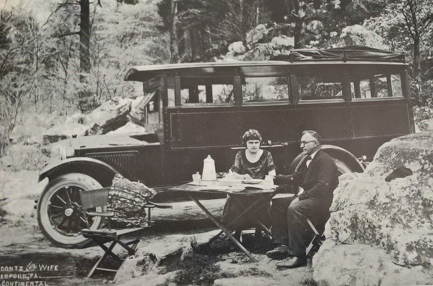 In 1925, Mr. and Mrs. S. H. Koontz share a meal outside of their parked house car, in Bedford. Before the spread of motels and motor courts, travelers would camp in or around their cars (Margolies).