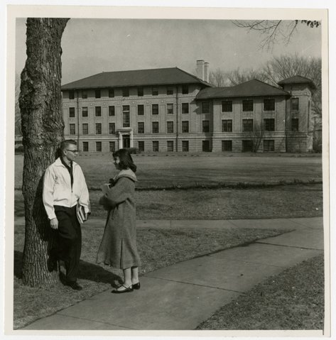 Two students standing beside a tree with Corwin and Larimore halls in the background. Photo courtesy of Elwyn B. Robinson Department of Special Collections, University of North Dakota. UAP28885.