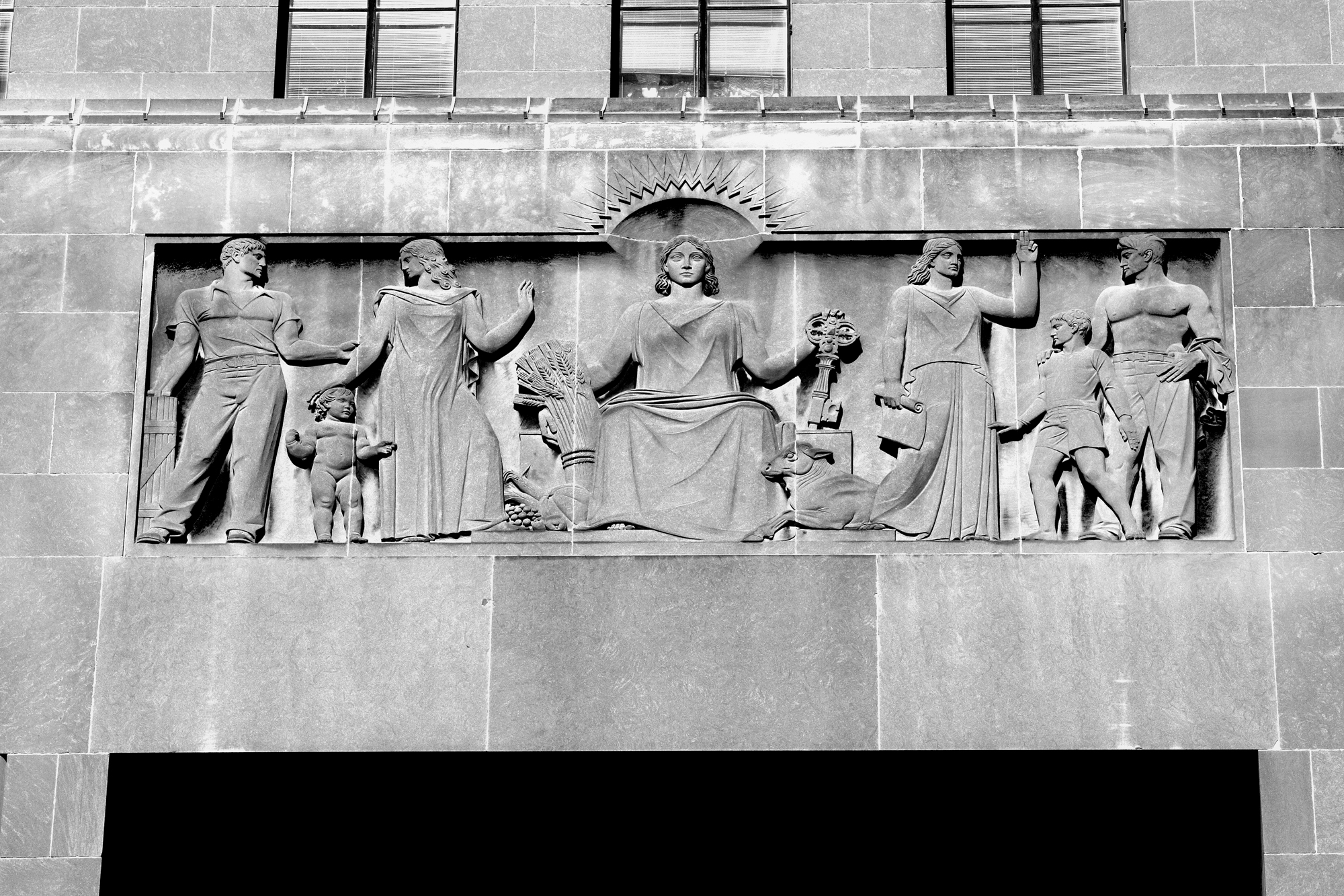 An image of the central panel of the 6th Floor Frieze on the South Face of City Hall. Center: A symbolic female figure representing Kansas City, seated, holding a sheaf of grain and the key to the city, surrounded by symbolic figures representing civic virtues. Left: Three figures representing Education with Law as its guardian. Right: Three figures, Faith holding the scroll of the future and Public Spirit represented by a young man and a boy.