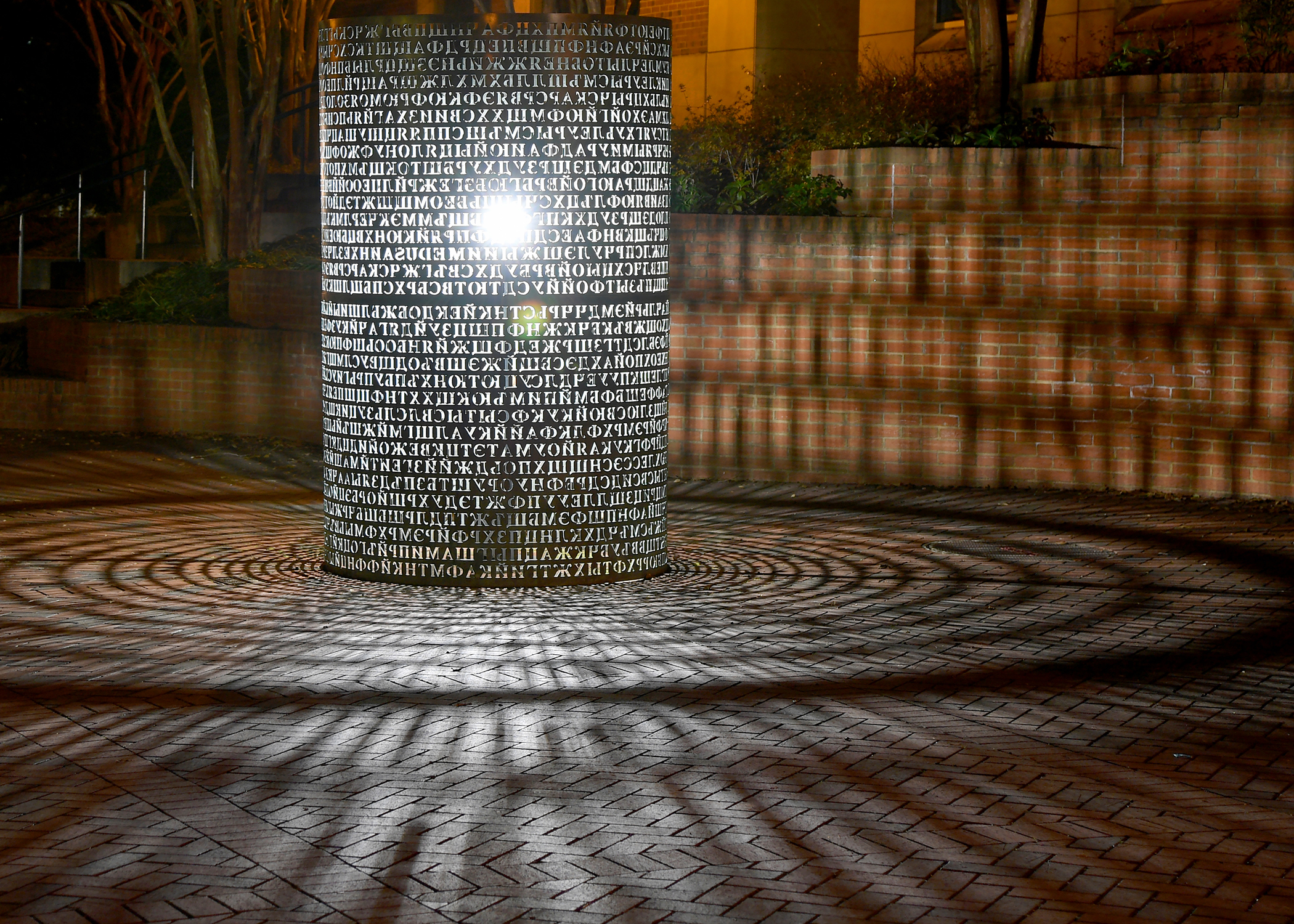 Bronze, cylindrical sculpture with Cyrillic letters cut out of the surface. Sculpture is illuminated from within at nighttime