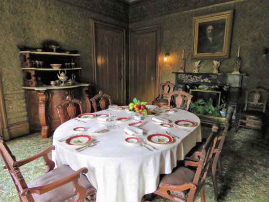 A shot of the dining room. The large, oval-shaped table is surrounded by six wooden chairs and covered with a white linen tablecloth. The table is set with red and white plates, and a fruit bowl serves as the centerpiece. 