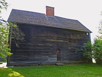 A shot of the Buttolph-Williams House. This wooden house is windowless and features a sloped roof, topped with a brick chimney.