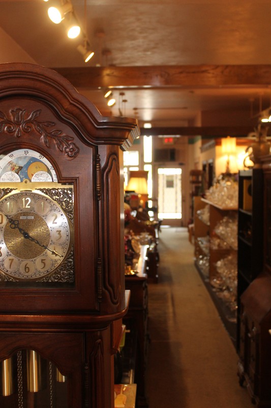 A bright showroom, with the glass rear door at center. The space is narrow and deep. Many glassware items are displayed for sale, and the face of a grandfather clock is in the left foreground..