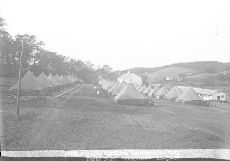 Black and white photo showing over 30 large canvas tents in the foreground, arranged neatly in three rows. Immediately behind the rows sits a large white barn, and below the last row of tents is a long white one-story structure. In the far distance another barn, and two houses sit on the hillside.