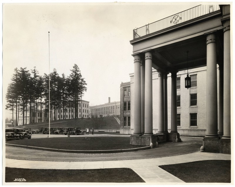 Black and white photograph depicts large columned entrance of Multnomah County Hospital to the right. In the center of the frame is a large driveway circle. In the background, a large classical style building (Mackenzie Hall) is visible. Several large evergreen trees are visible to the left.