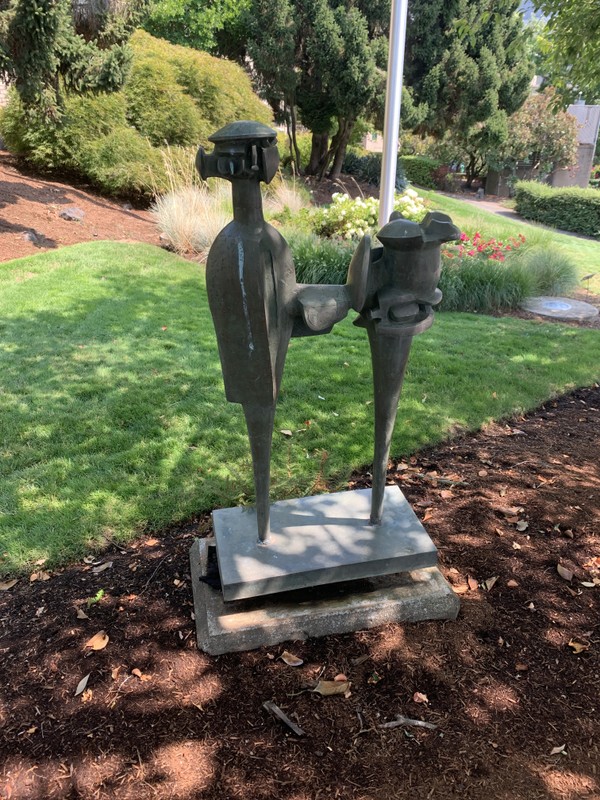 A color photograph depicts a bronze, 47” x 24” x 12.5” modernist statue of a figure holding a geometric object. In the background, lawn and shrubbery is visible