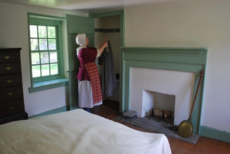 A woman dressed in period clothing hangs up a petticoat in a small closet next to the fireplace. A low-post bed is in the foreground.