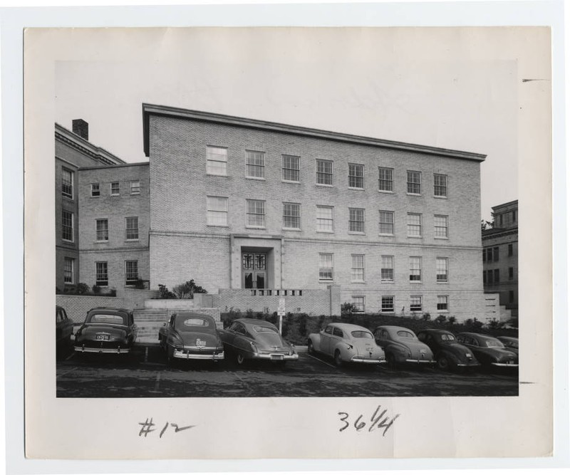 Black and white photograph of a four-story brick building with early 1950s style cars visible in the foreground.