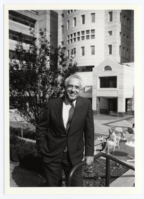 Black and white photograph of Leonard Laster, M.D., OHSU President (1978-1987), in courtyard outside Vollum Institute for Biomedical Research. Laster wears a dark suit and white tie, and smiles in front of a courtyard and a tall curved building.