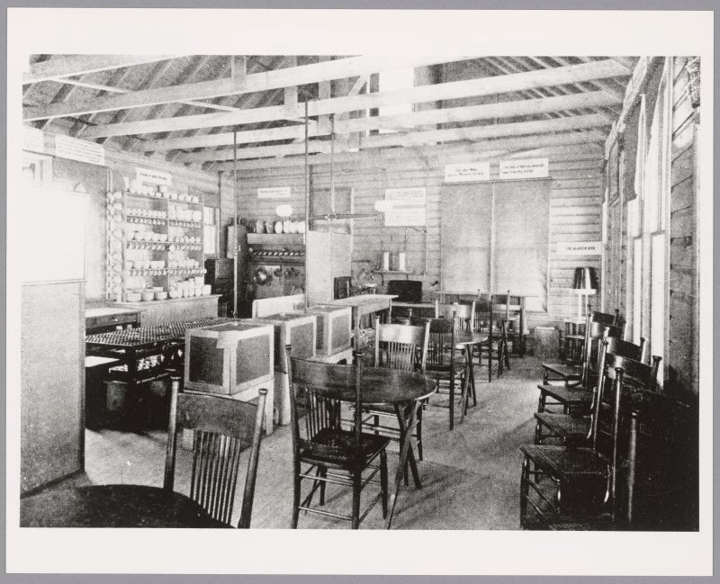 Lunchroom with small dark tables with two chairs each. Shelving with dishes and kitchen tools line the walls.