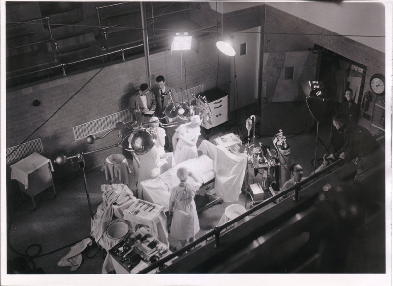 Black and white photograph of the Multnomah County Hospital surgery theater during a filming event put together by the Oregon State System of Higher Education (OSSHE), showing an operating team in white seen from above operating on a patient whose face is not visible.