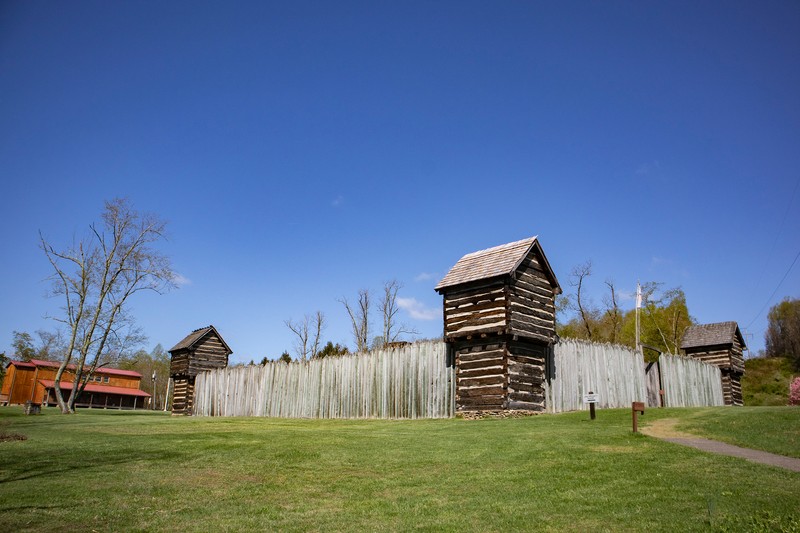 Pricketts Fort is shown with the log stockade walls, three out of four corner blockhouses visible, and the visitors center in the lefthand corner. The stockade walls are pointed at the top. The blockhouses are built with logs and clay chinking in between; they have a gabled roof and small lookout windows. The sky is bright blue.