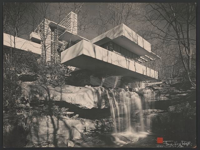 This black and white photo depicts the scenic exterior of Fallingwater. The home sits atop a waterfall that flows underneath, the home is surrounded by the dense woods within the Laurel Highland.