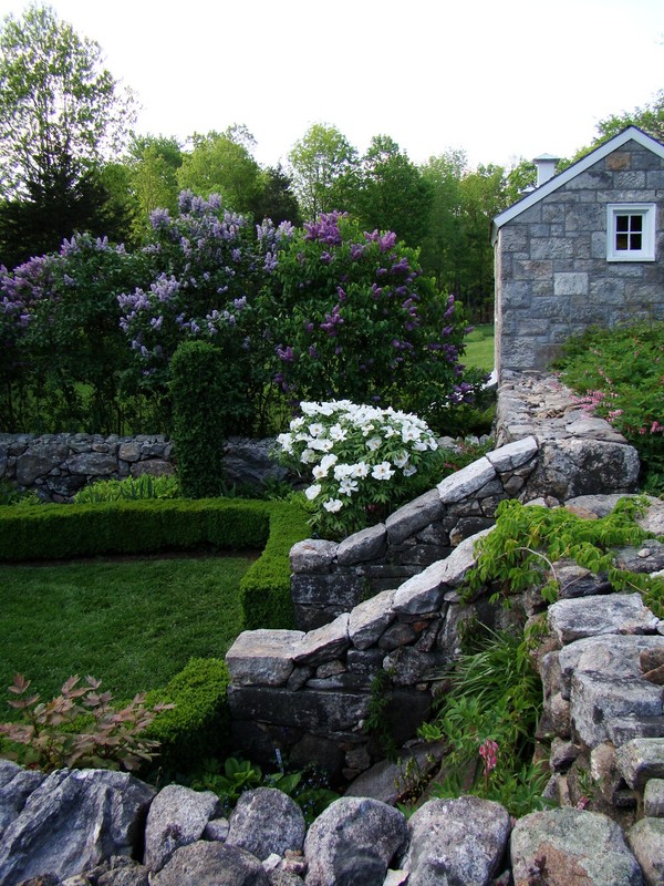 A shot of the gardens at Weir Farm. A low stone wall runs around the perimeter of the garden. Stone steps lead down into the space. A stone cottage can be seen in the distance. Several flowering shrubs are present.