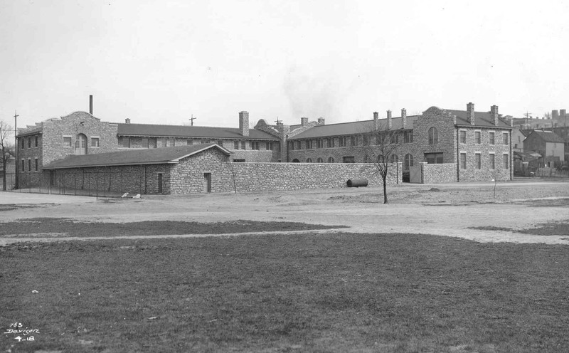  A picture of a two-story Mission Revival -style limestone building. The building has a courtyard in the front, surrounded by a stone wall. In the foreground, a large open grassy area is visible.