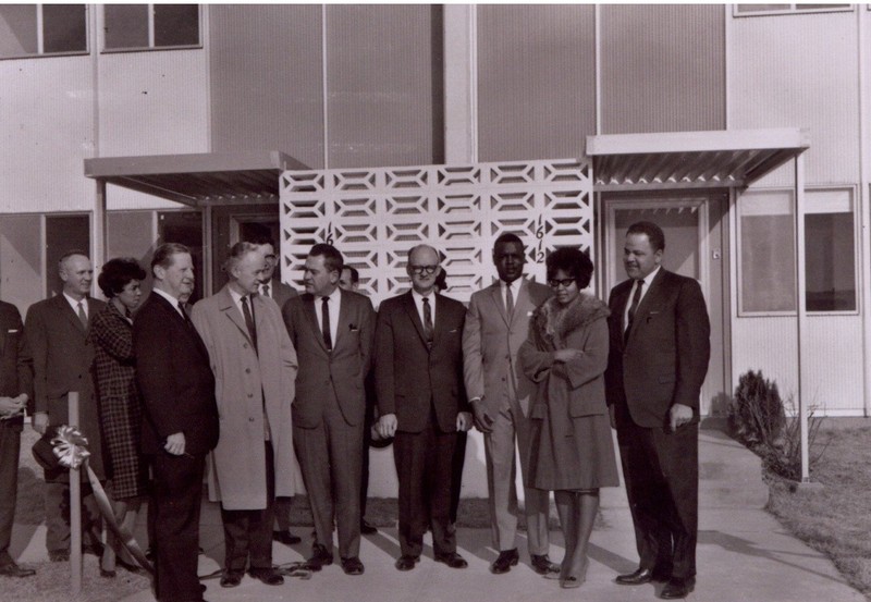The image shows a group of thirteen people gathered to take a picture in front of two adjoined townhouses. The leftmost and second from left people are white men in dark suits, standing and watching the proceedings. The third person from the right is an African-American woman in a dark-colored checkered dress, watching the proceedings. The next seven people from the left are all white men in suits. Four of them are standing in a line, as if posing for the picture, while three of them are obscured behind the others. The three rightmost people are African-American. The third person from the right is an African-American man in a medium-colored suit. The second person from right is an African-American woman in a medium-colored coat with a fur collar. The rightmost person is an African-American man in a dark suit, identified in supplementary documents as Bruce R. Watkins.