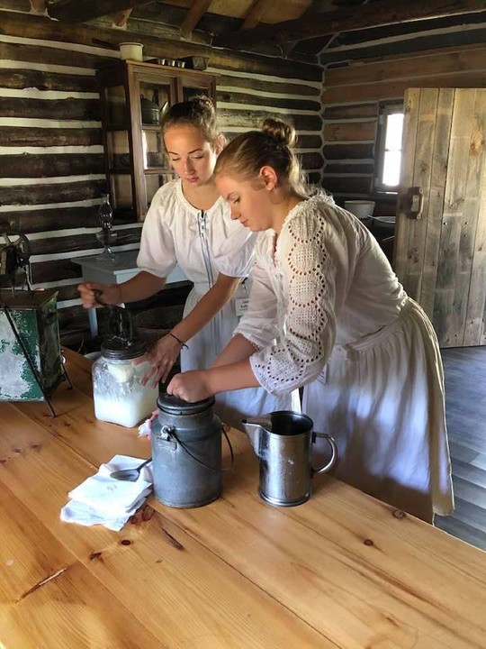 Volunteers (sisters) making butter in a traditional-style home, July 2020. The COVID-19 pandemic provided some challenges to the Pioneer Village Museum during 2020, but with many volunteers working together as families, some of the challenges were able to be overcome.
