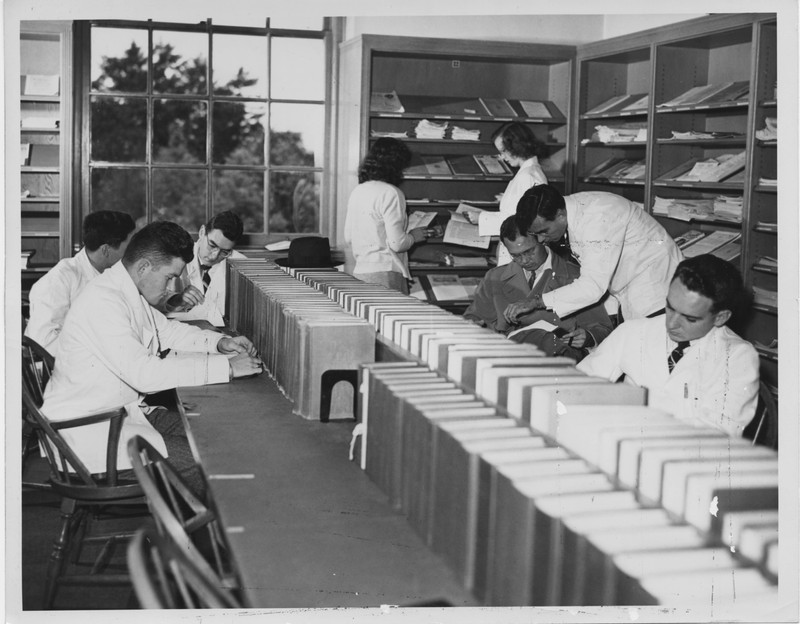 Black and white photograph of reference activities at the Old Library, including students sitting at a long table lined with books, and two students standing and looking at books next to a shelf.