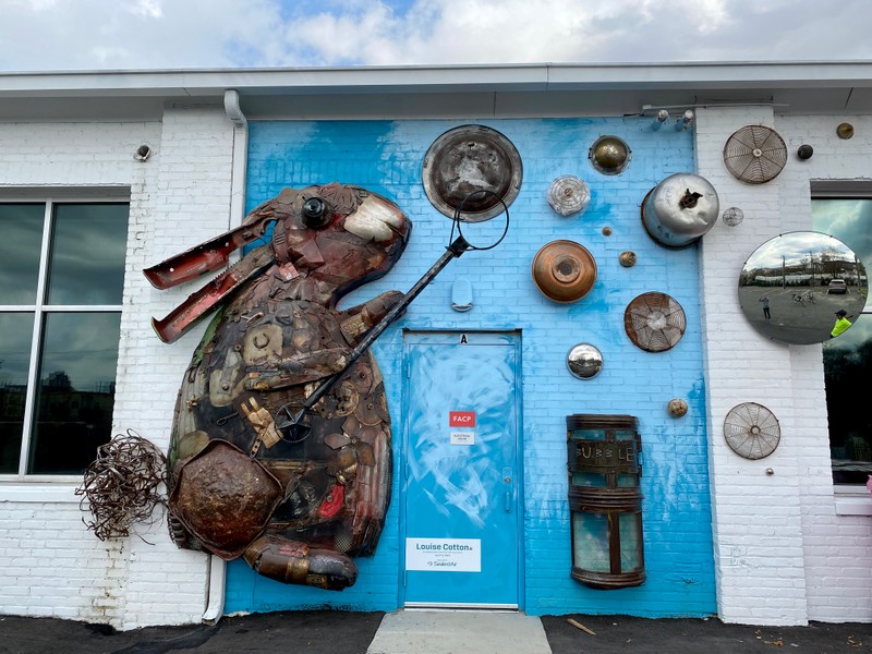 A large bunny made of recycled materials waves a bubble wand and it attached to a blue wall.