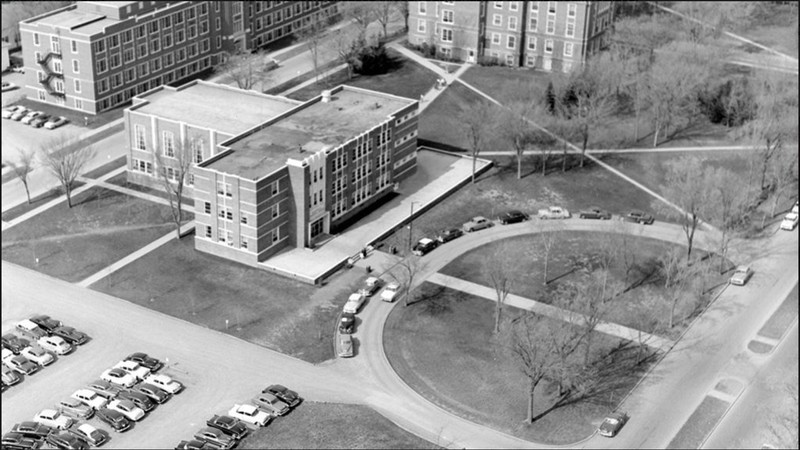 Black and white aerial image of a building, with cars in a parking lot next to it