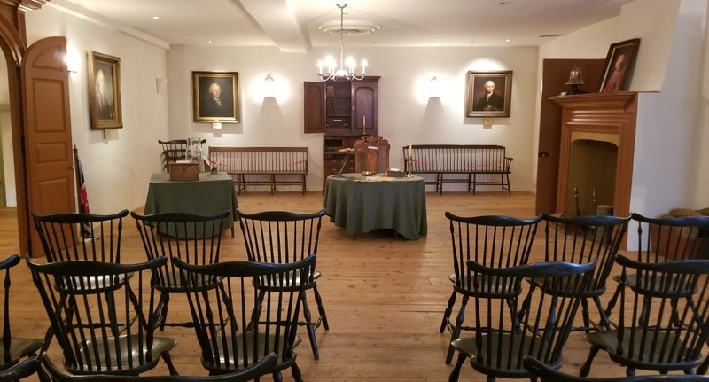 Room with rows of black windsor chairs, large doorway to the left, two tables in the background and portraits along the walls. 