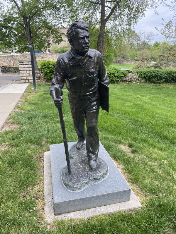 A statue of Benton. He is holding a sketch book and waling with a walking stick.