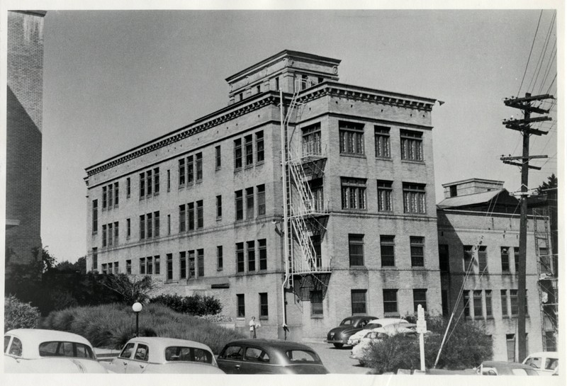 Black and white photograph of the exterior of Doernbecher Memorial Hospital for Children with cars visible in the foreground. The viewpoint is from a parking lot on the southest side looking northeast.