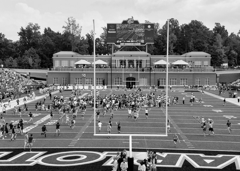 The football field with view of both field goals and scoreboard. Players and other people are running towards the field. 