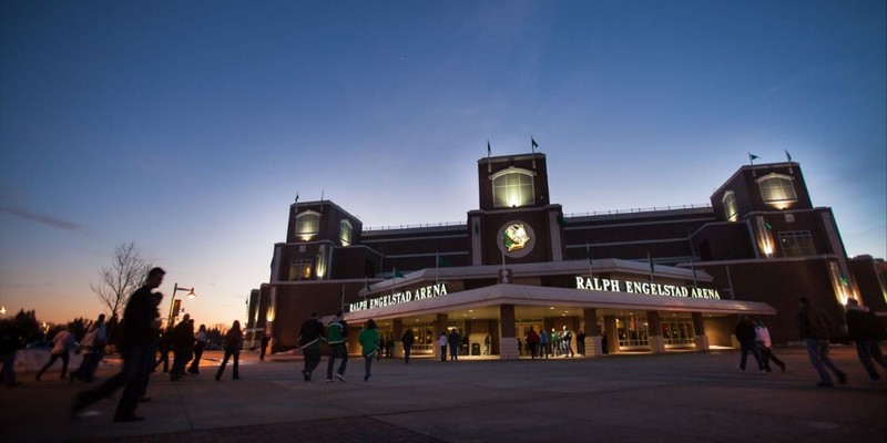 People approach the red brick arena main entrance at dusk. Lights illuminate the text "Ralph Engelstad Arena" and a Sioux head logo.