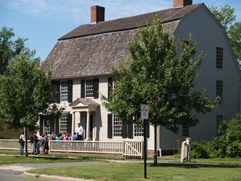 A side view of the Joseph Webb House. The two-story home features white siding and black shutters. A narrow porch runs along the front of the house. A small tree obstructs part of the view of the house.