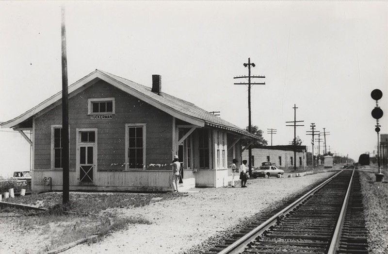 A black and white photo a building with people standing outside of it in front of some train tracks.
