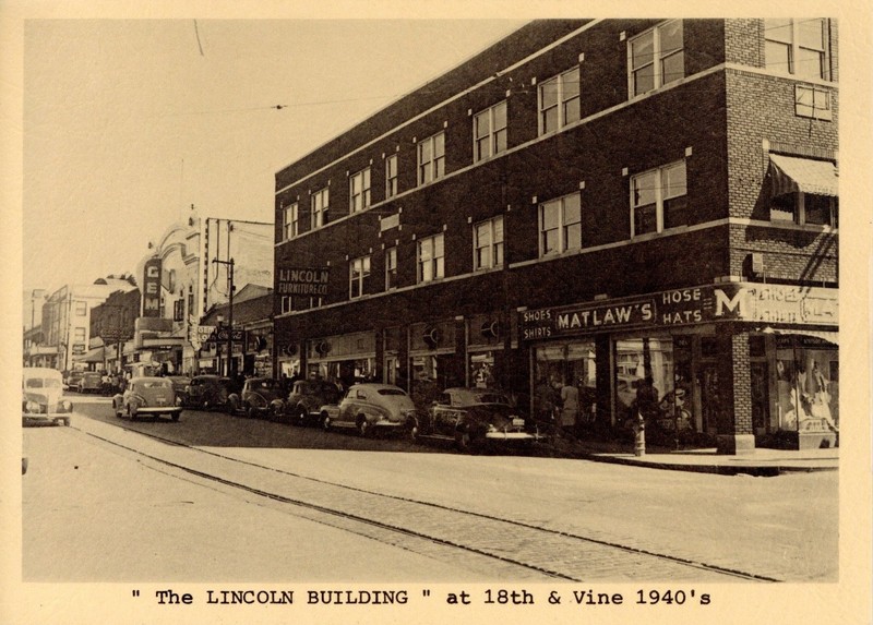A picture of a three-story brick building. On the right side of the image, there is a storefront on the corner of the building with a sign above it reading "MATLAW'S SHOES SHIRT HOSE HATS". To the left of the corner storefront, additional storefronts can be seen. The second and third floor of the building have rows of windows. Along the sidewalk in front of the building, several cars are parked. In the background, additional buildings are visible, including the Gem Theater. In the foreground, trolley tracks are visible, set into the pavement of the street.