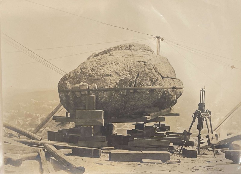 A black and white image of the boulder elevated with wooden blocks and held in place with a large metal brace.  There is equipment in the foreground and in the background there are electrical wires.  